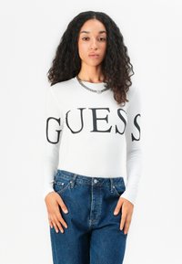 Long-sleeve, fitted white top with "GUESS" in bold black letters, paired with high-waisted blue denim jeans featuring a classic button closure.