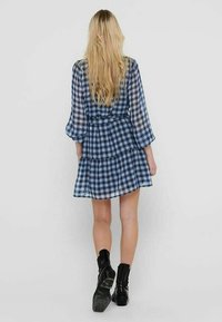 Blau-weißer kariert Kleid mit gestuften Rüschen und langen, transparenten Ärmeln, kombiniert mit knielangen schwarzen Stiefeln.