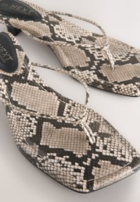 Sandales plates avec un motif peau de serpent, présentant une texture tissée en beige et noir, un design à bout carré et des brides minimalistes.