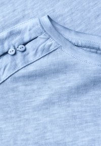 Camicia di cotone azzurro chiaro con scollatura rotonda, caratterizzata da due bottoni decorativi sulla spalla e un sottile motivo testurizzato.