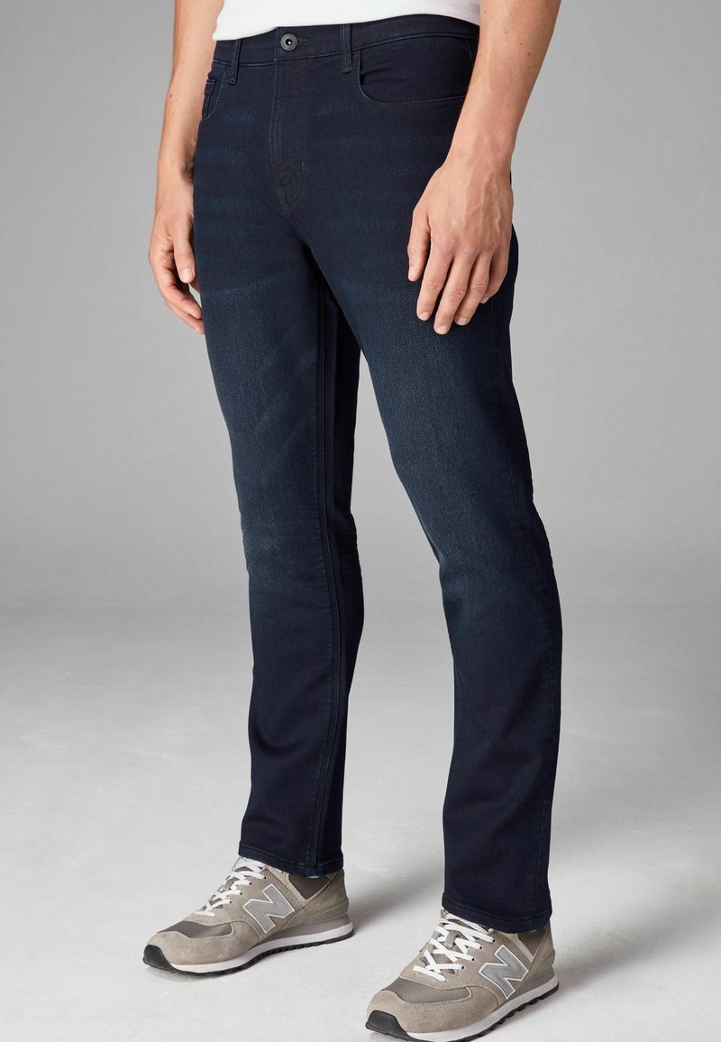 Jeans de mezclilla azul oscuro con un ajuste Slim; cuenta con cinco bolsillos, cierre de botón y sutiles rayas verticales. Combinados con zapatillas deportivas grises.