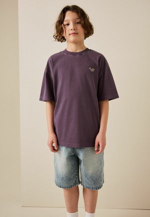 Göndör hajú gyermek, laza lila pólót és világoskék farmer rövidnadrágot visel, egyszerű bézs háttér előtt áll.