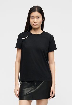 Hosszú sötét hajú nő fekete pólót visel, amelyen fehér szárny alakú kitűző van a vállán, valamint fekete bőr miniszoknyát, egyszínű háttér előtt.