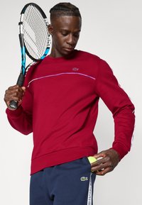 Bordeaux genser med en kontrasterende lilla stripe, med Lacoste-logo. Mørkeblå bukser, holder en gul tennisball i lommen.