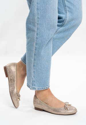 Beige metallische Ballerinas mit kleinem Schleifchen und Perlenakzenten, getragen zu hellblauen, gekürzten Jeans bei gekreuzten Füßen.