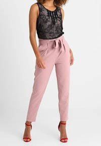 Černý krajkový top na ramínka s kulatým výstřihem, doplněný o růžové kalhoty s vysokým pasem a vázankou. Celkový vzhled doplňují červené boty na podpatku.