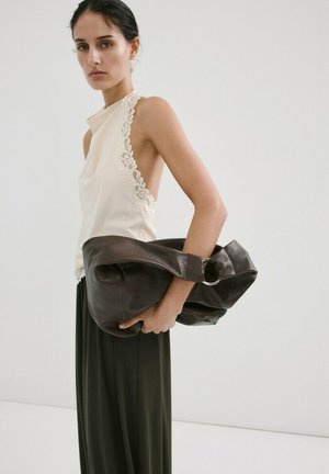 Femeie purtând un top alb fără mâneci, cu dantelă, și o fustă neagră, ținând o geantă mare din piele neagră, pe un fundal simplu.