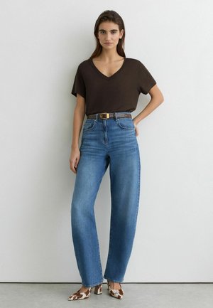 Žena stojaca opretá o bielu stenu, oblečená v tmavohnedom tričku s V-výstrihom, modrých vysokoplnených džínsach s opaskom a na podpätkoch s potlačou kravy.