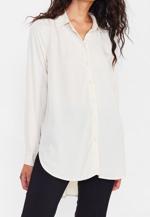 Cream-colored long-sleeve shirt with a button-up front, featuring a subtle textured pattern and rounded hem. Fabric has a smooth finish.