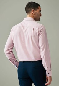 Helles rosa Langarmhemd mit gemusterter Textur und kontrastierenden blauen Manschetten, kombiniert mit dunkelblauen Hosen. Zeigt die Rückansicht und das Kragendesign.
