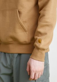 Brown hoodie with a front pocket and ribbed cuffs, featuring a small orange logo on the left sleeve. Smooth fabric texture.