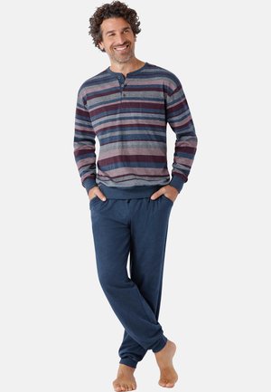 Mann steht barfuß, trägt ein blau-bordeaux gestreiftes Langarmshirt und einfarbige blaue Jogginghose mit Händen in den Taschen.