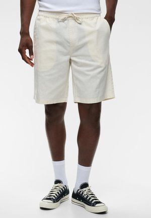 Homme portant un short écru à cordon, des chaussettes blanches montantes et des baskets basses noires, debout les mains dans les poches.