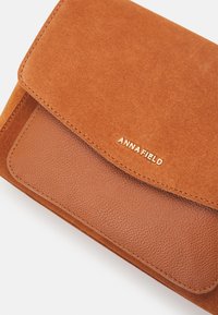 Brun læderhåndtaske med en glat nederste del og en øverste flap i ruskind, med et guldpræget logo.