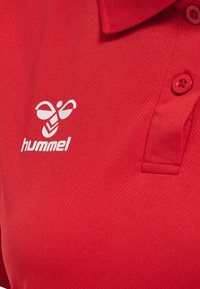 Rød polo shirt lavet af tekstureret stof, med et hvidt logo og tre knapper med runde røde detaljer på kraven.