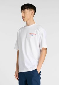 Valkoinen puuvillapaita lyhyillä hihoilla, jossa on punainen ja sininen "new balance" -logo vasemmassa rinnassa. Rentomallinen, pyöreä kaula-aukko.