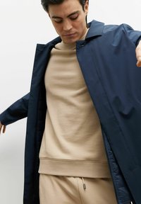 Granatowa kurtka zewnętrzna o minimalistycznym designie, z kołnierzykiem i gładką teksturą, noszona na beżowym swetrze.