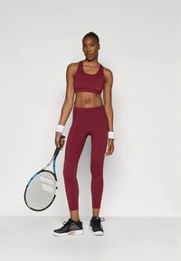 Tumši sarkans sporta krūšturis un legingi ar piegulošu dizainu. Modelis tur tenisa raketi, valkājot melnas sporta kurpes un baltas aproces uz rokām.