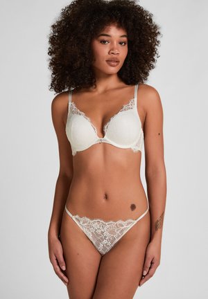 Vrouw met krullend haar die een witte kanten beha en bijpassende kanten slip draagt, staand tegen een effen lichtgrijze achtergrond.