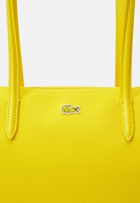Sac fourre-tout jaune en matière texturée. Comprend deux poignées et un petit logo crocodile brodé. Surface lisse avec des détails cousus.