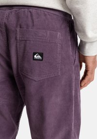 Lilla corduroy-bukser med baklomme og et svart logo-patch, som viser ribbet tekstur og en avslappet passform.