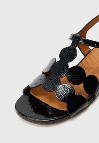 Melna lakotas ādas sandāle ar atvērtu purngalu un viļņotu dizainu. Brūna zolīte ar redzamu faktūru un veidotu formu.