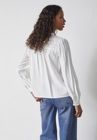 Hvid langærmet bluse med broderet detalje på rygstykket, parret med lyseblå løstsiddende jeans. Glat stofstruktur.