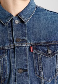 Modrá džínová bunda s klasickým designem, vybavená kovovými knoflíky, dvěma náprsními kapsami a malou červenou značkou na levé kapse.