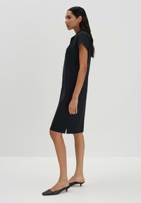 Robe noire longueur genoux avec une coupe décontractée, des manches courtes et des fentes sur les côtés. Réalisée en tissu lisse, assortie à des mules noires à bout pointu.