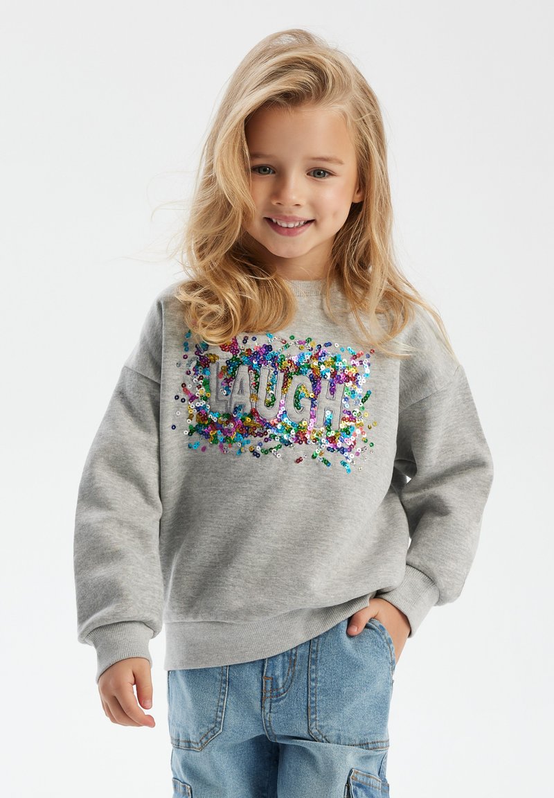 Felpa grigia con una grafica "LAUGH" realizzata in paillettes colorate, caratterizzata da una vestibilità comoda e polsini a costine. Abbinata a pantaloni cargo blu chiaro.