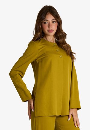 Donna con lunghi capelli castani che indossa una maglia gialla senape a maniche lunghe con bottoni e pantaloni coordinati, che guarda leggermente di lato.