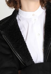 Chaqueta negra con un cuello de felpa sobre una camisa blanca de botones. La chaqueta tiene un frente con cremallera y una textura suave.