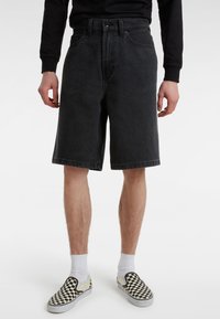 Svarta denimshorts för män med lös passform, midjehöjd och fransad fåll. Kombinerade med svartvita rutiga slip-on-skor och vita strumpor.