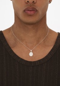 Silberne Anhänger-Kette mit einem ovalen Charm, der ein graviertes Design zeigt, an einer zarten Kette, getragen über einem strukturierten dunkelbraunen Oberteil.
