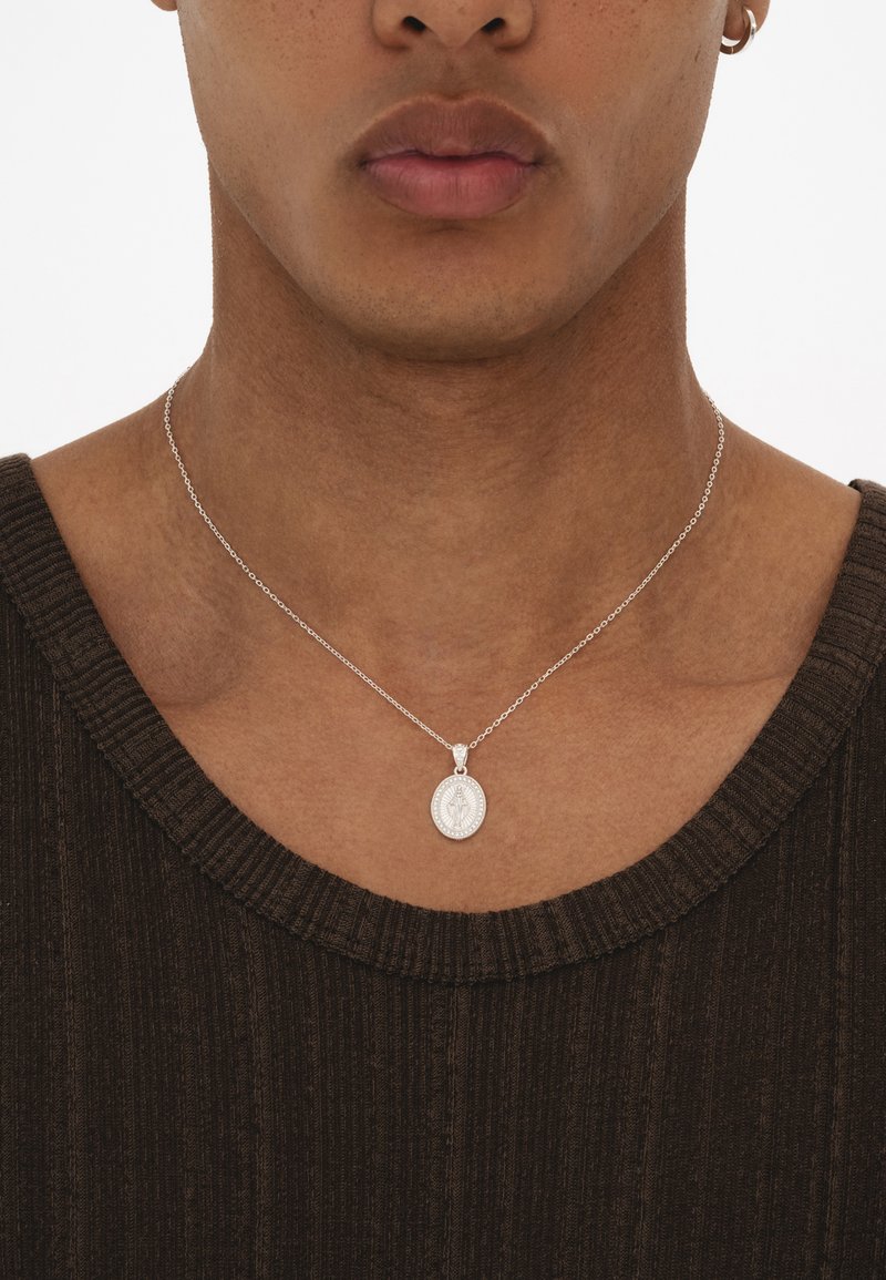 Silberne Anhänger-Kette mit einem ovalen Charm, der ein graviertes Design zeigt, an einer zarten Kette, getragen über einem strukturierten dunkelbraunen Oberteil.