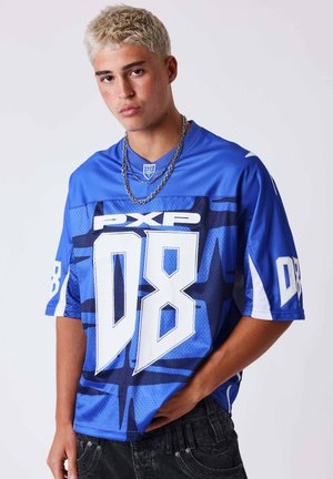 Giovane uomo con capelli corti decolorati che indossa una maglia sportiva blu con testo bianco "08" e "PXP", e jeans neri, su sfondo bianco.