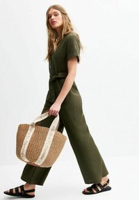Olive-green jumpsuit with short sleeves and a belted waist, paired with a woven straw tote featuring beige straps with printed text, and black sandals.