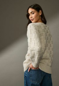 Pull en tricot couleur crème avec un motif à carreaux texturé, col rond et ourlet côtelé, associé à un jean en denim bleu.
