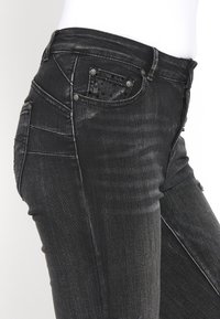 Gros plan sur un jean en denim noir avec poches avant et arrière, passants de ceinture et texture subtilement délavée sur un mannequin blanc.