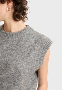 Grauer ärmelloser Strickpullover mit einem gerippten Halsausschnitt und Armausschnitten. Die weiche Textur und subtile Farbvariationen sind überall sichtbar.
