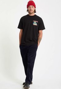 Must puuvillane t-särk, millel on värvikas graafiline plaastr vasakul rinnal, kombineeritud tumeliste vööpüksidega ja mustade kingadega. Peal punane kootud müts.