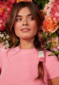 Pink ribbed T-shirt, shoulder-length dark hair with pastel scrunchies in green, white, and pink, surrounded by a floral backdrop.