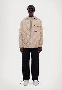 Giacca beige trapuntata con tasca anteriore, maglione grigio, pantaloni blu e scarpe da ginnastica bianche. Gli accessori includono un cappello nero e occhiali da sole.