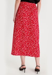Jupe midi rouge à fleurs avec taille smockée, ornée de marguerites blanches et d'accents verts. Tissu lisse, associée à des sandales à talons noires.