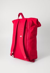 Red fabric backpack with adjustable straps, top handle, mesh panel, and zippered side pocket. White logo on front. Soft, structured design.
