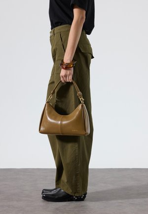 Persona con pantalones color oliva y zapatos negros sosteniendo un bolso de cuero marrón con pulseras de carey en la muñeca, de pie sobre un suelo gris.