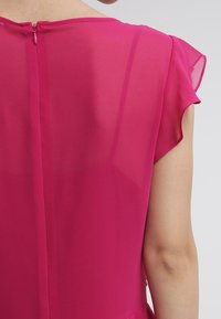 Blusa fucsia con una tela ligera y transparente, mangas cortas con volante y cierre de cremallera en la parte posterior con un pequeño botón de lazo.