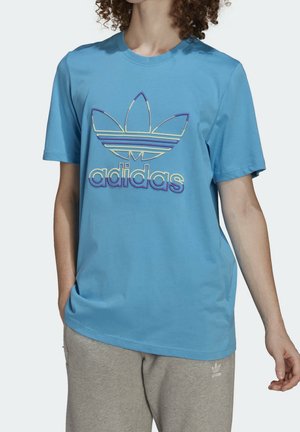 Person trägt ein blaues Adidas-T-Shirt mit mehrfarbigem Trefoil-Logo und grauer Adidas-Sweathose, steht mit einer Hand in der Tasche.