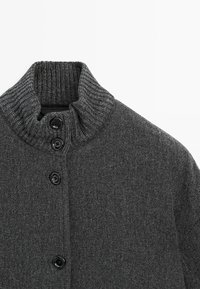 Pull en maille gris avec un col haut à côtes, trois boutons noirs et un tissu texturé. La vue en gros plan met en avant les détails du matériau.