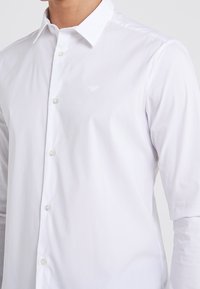 Bílá košile na knoflíky s límečkem, dlouhými rukávy a malým vyšitým logem na hrudi. Vyrobena z hladké tkaniny s čistým zakončením.